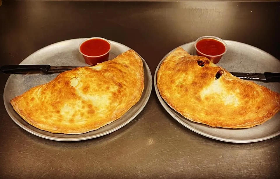 Two golden calzones on pizza pans, each with a cup of marinara